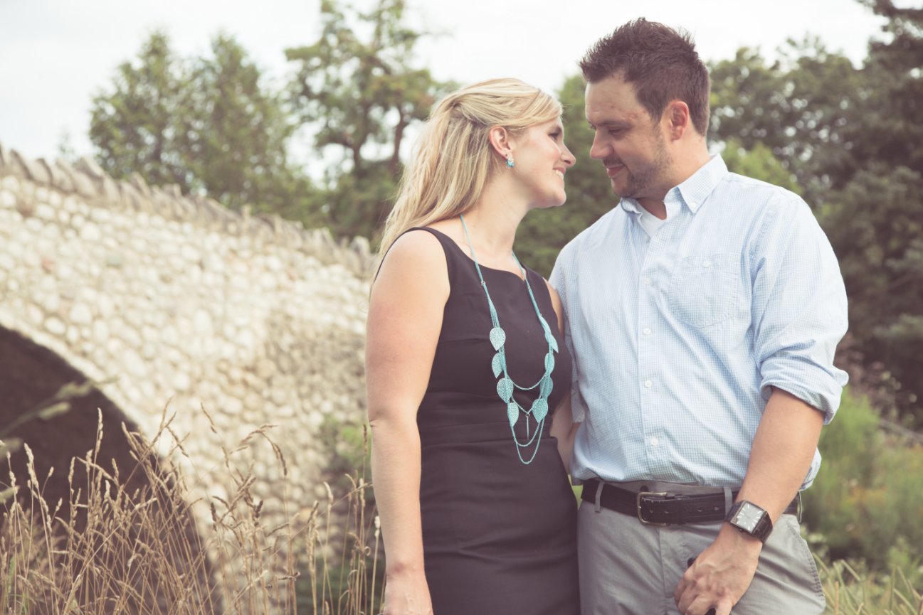 A couple smiles at each other near a stone bridge with greenery around them. Websters Falls