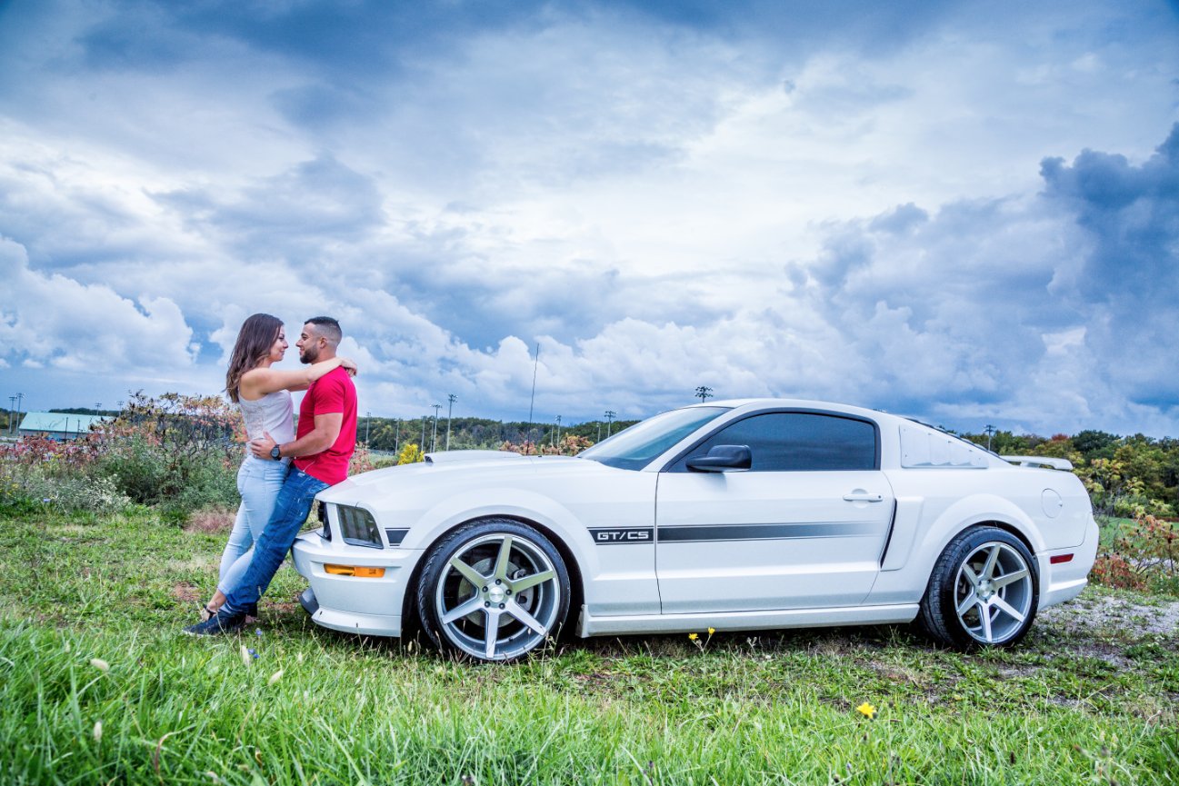 A couple embracing near a white sports car on a grassy field under a cloudy sky.