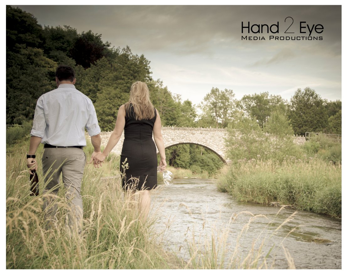 A couple walks hand in hand along a riverside path, facing a stone bridge, surrounded by lush greenery. Websters Falls