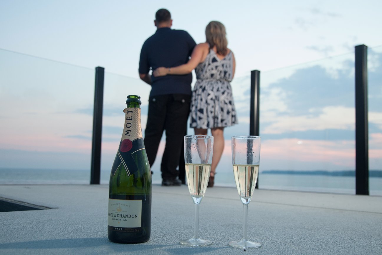 Couple embracing on a terrace at sunset, with champagne bottle and two filled glasses in the foreground.