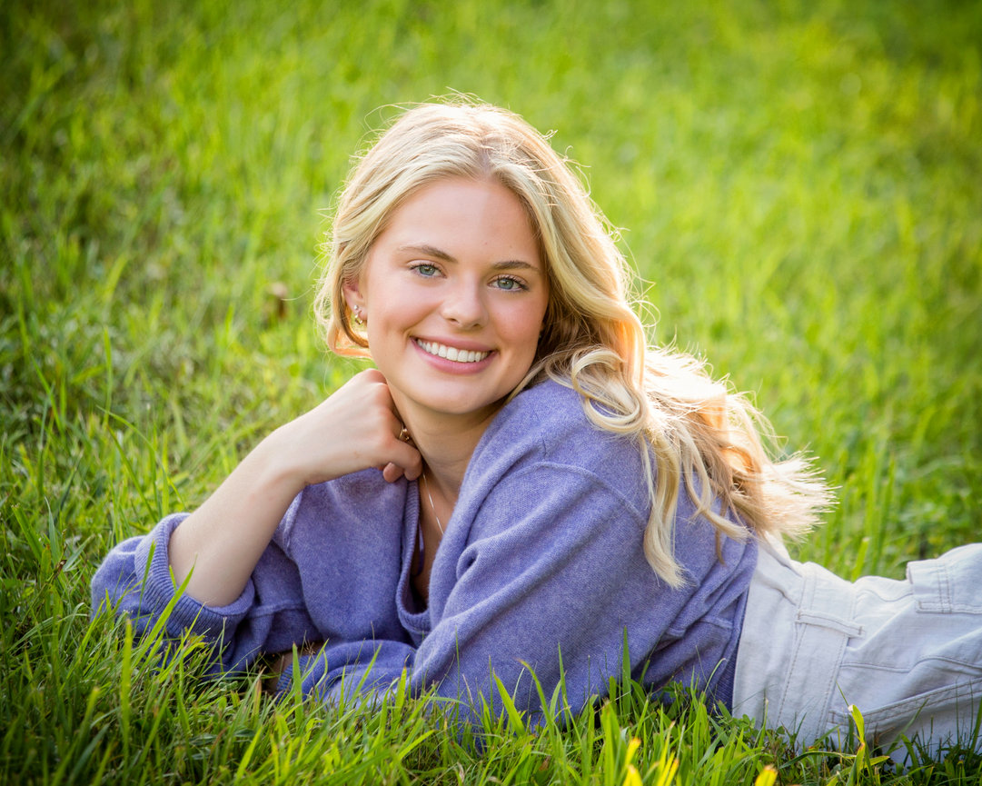 A person with long blonde hair smiling, lying on grass wearing a purple sweater.
