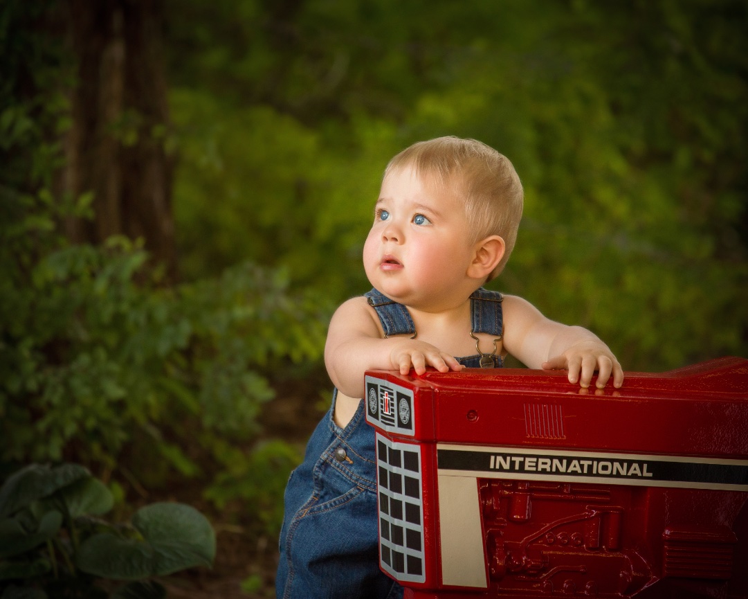 A baby in denim overalls leans on a red toy tractor outdoors with a forest background.