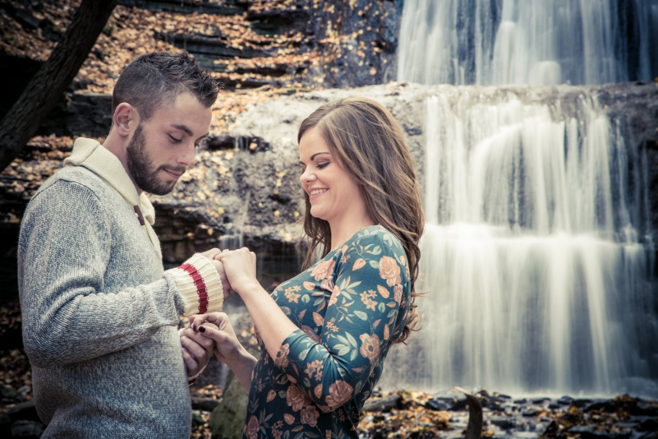 A couple smiling and holding hands in front of a waterfall, surrounded by autumn leaves. Sherman Falls, Ancaster, On