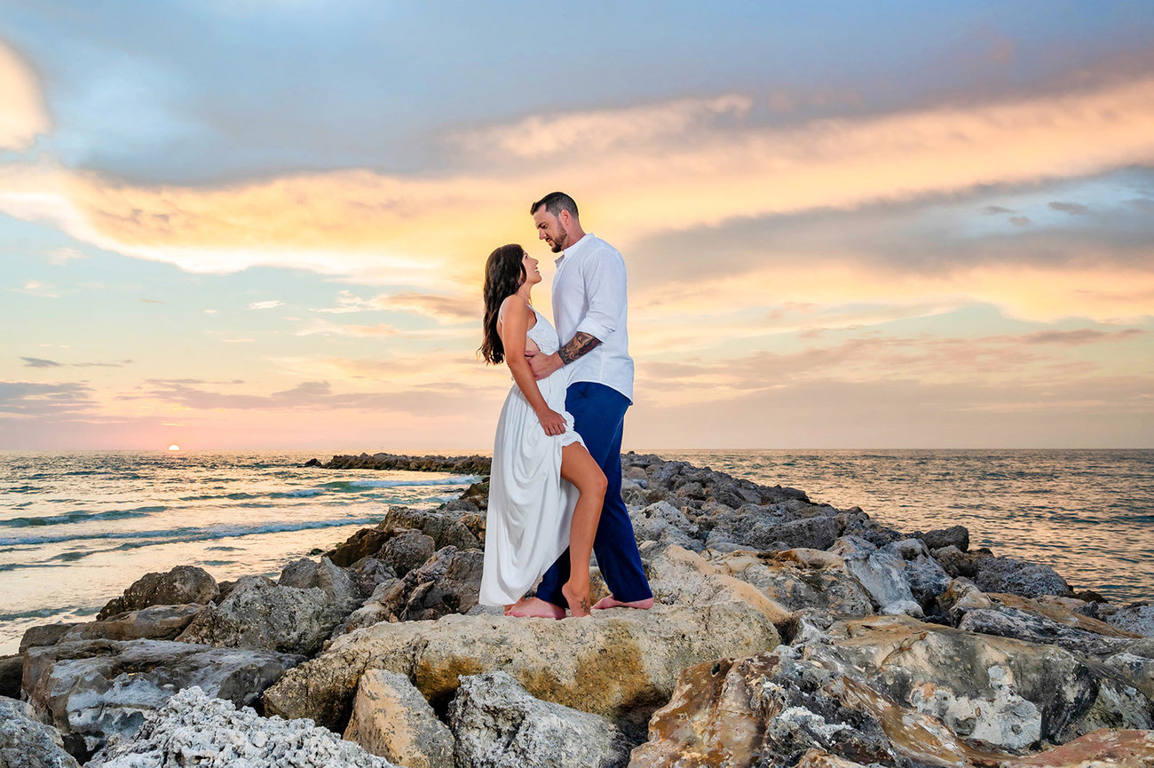 Couple in love standing on rocky shoreline at sunset, with colorful sky and gentle waves in the background.