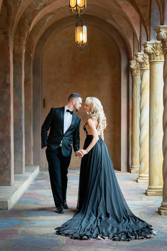 Elegant couple in formal attire holding hands in a stunning arched corridor with soft lighting.