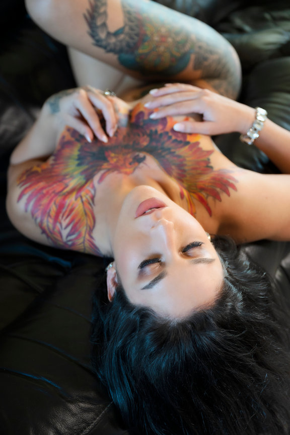 A woman reclining on a leather couch, showcasing a vibrant floral tattoo on her chest and arms.