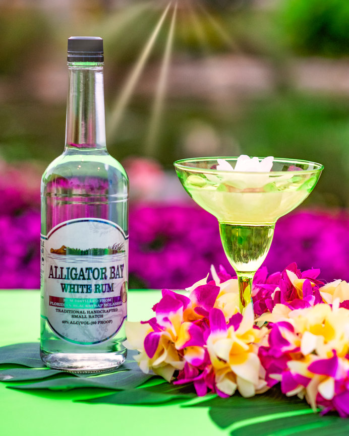 Bottle of Alligator Bay White Rum with a cocktail glass and floral decorations, set against a vibrant background.