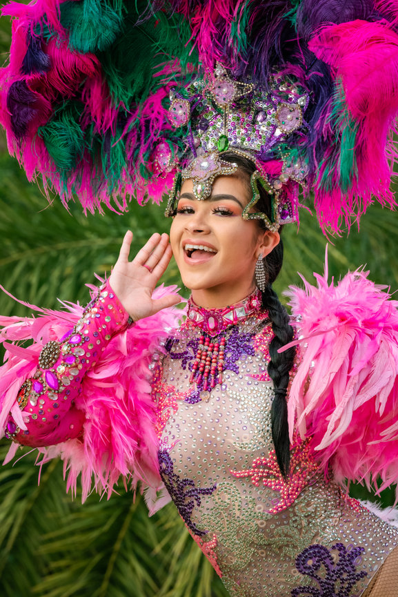 Young performer in vibrant pink feather costume with glittering accessories, joyfully posing against a green backdrop.
