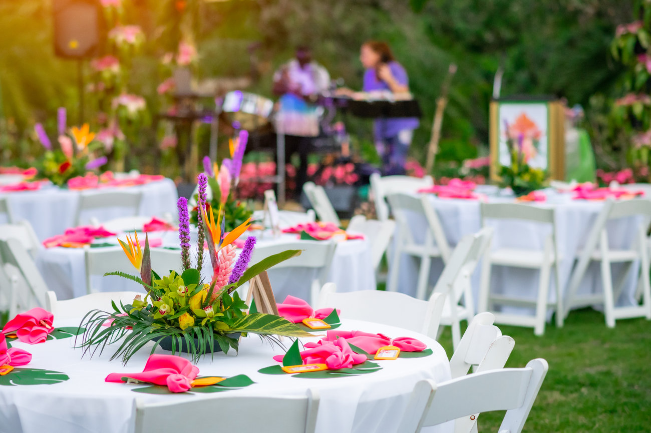 Colorful tables set with pink napkins and tropical centerpieces at an outdoor event, with musicians in the background.