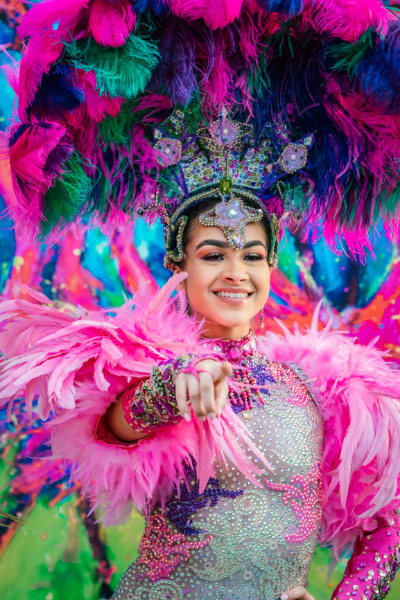 Smiling performer in vibrant, feathered costume with a colorful headdress at a carnival or festival.