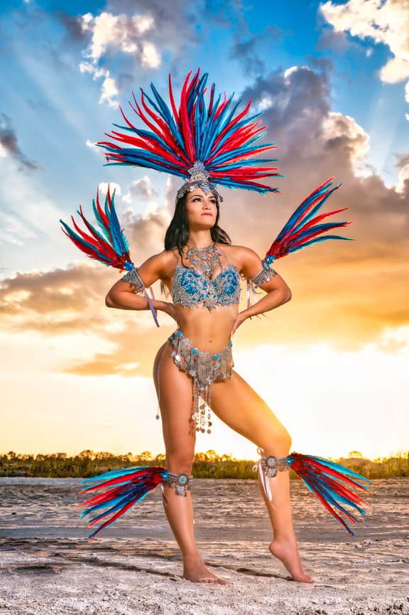 A woman in a colorful feathered costume poses confidently against a vibrant sunset.
