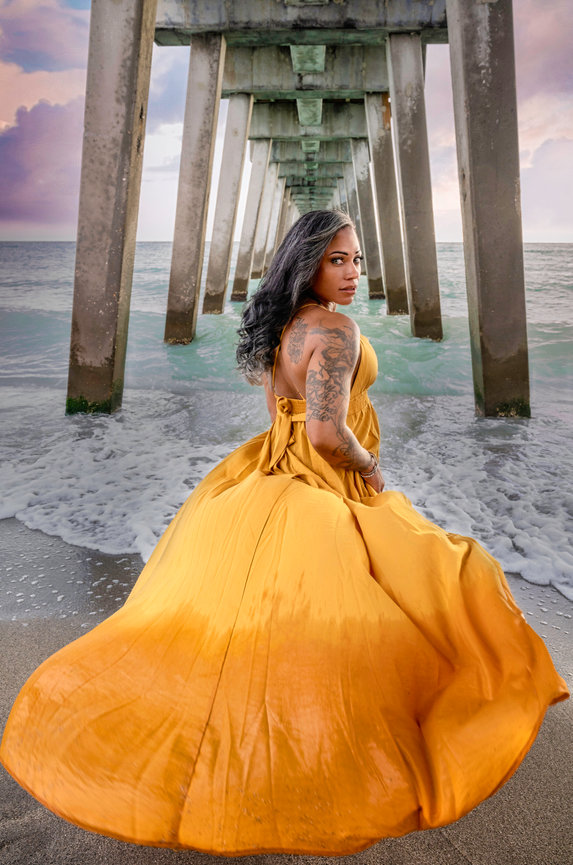 Woman in a flowing yellow dress stands under a pier, with ocean waves and a sunset backdrop.