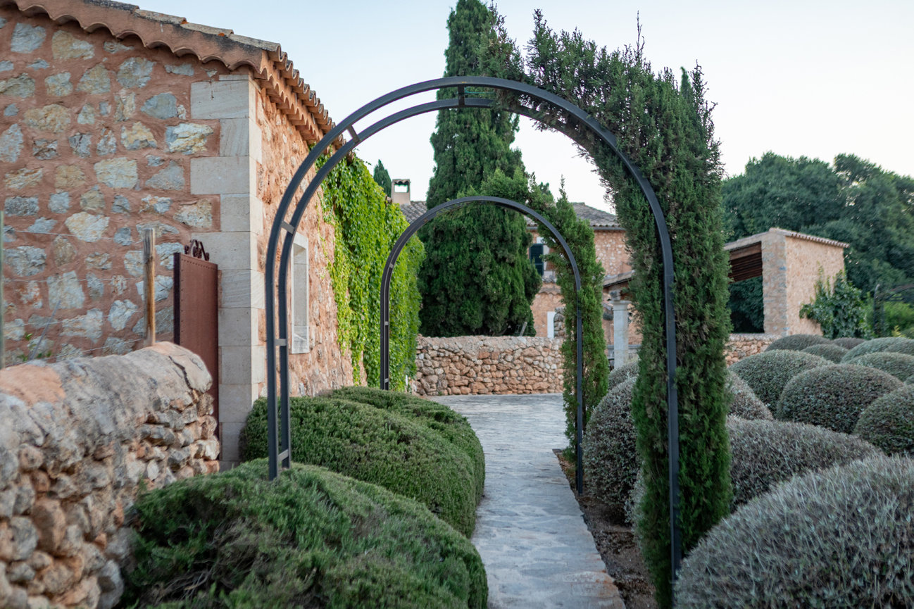 Stone pathway with metal arches, surrounded by greenery and Mediterranean-style stone buildings.