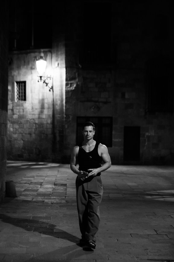 A man in a tank top and pants walks alone on a dimly lit cobblestone street at night. Barcelona night