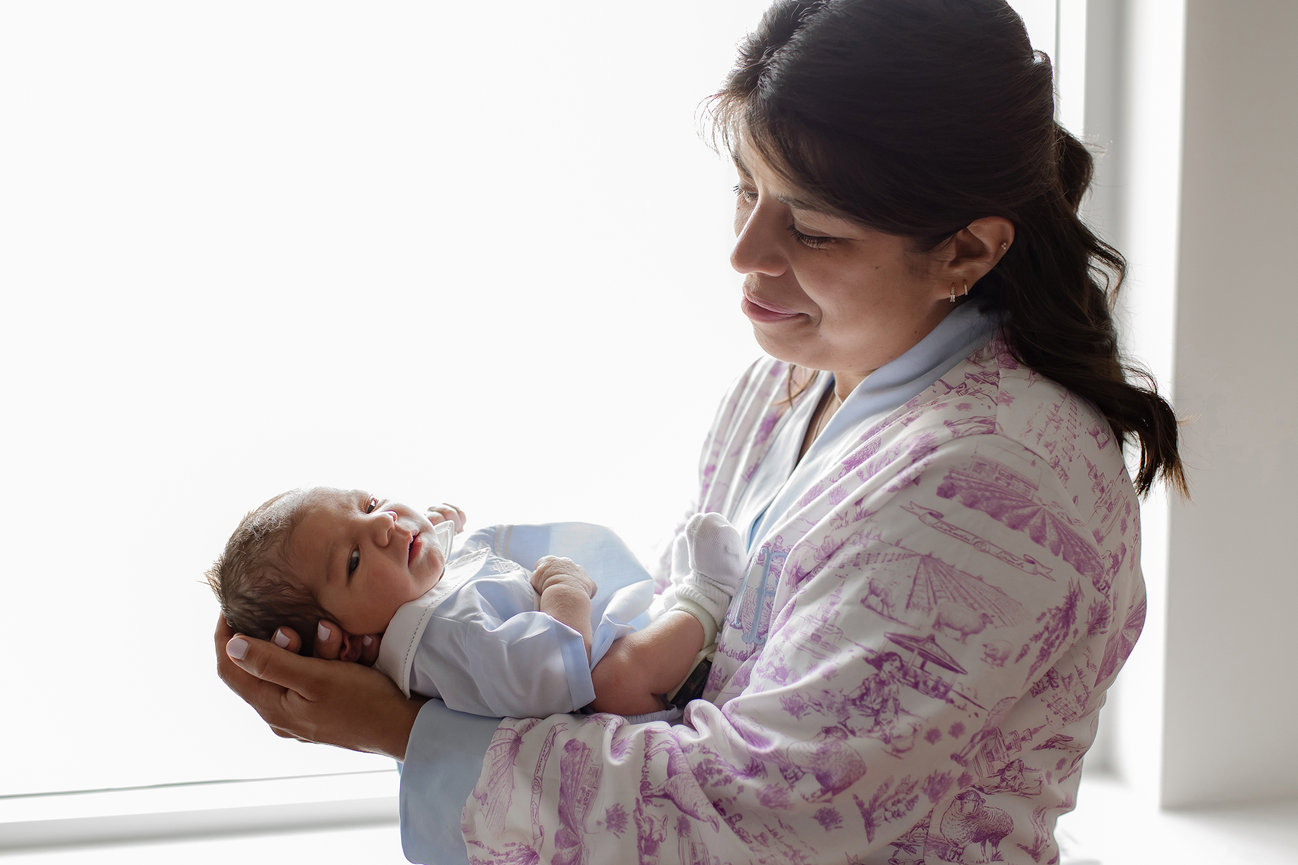 A woman gently cradles a newborn near a sunlit window.