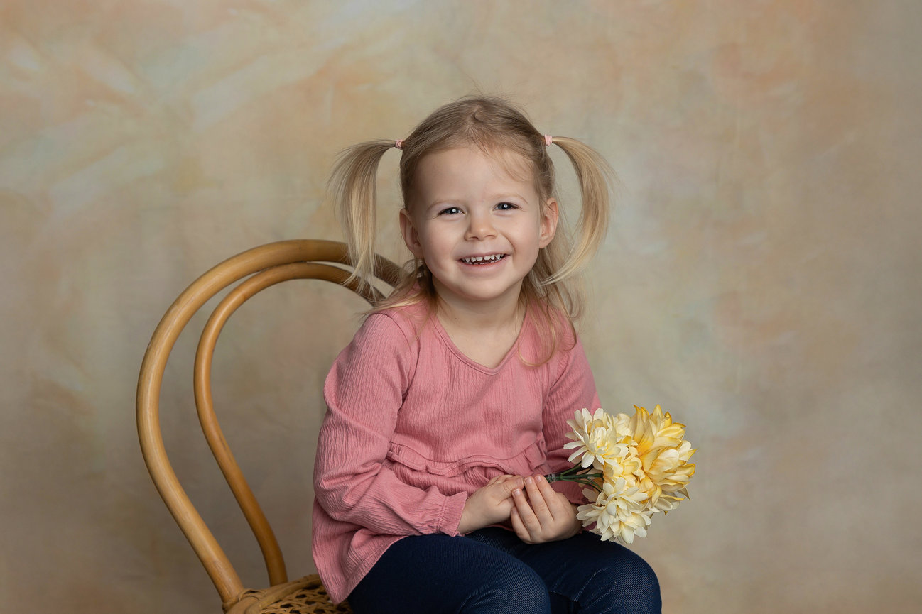 Young girl sitting on a chair, smiling, holding yellow flowers against a soft beige background.