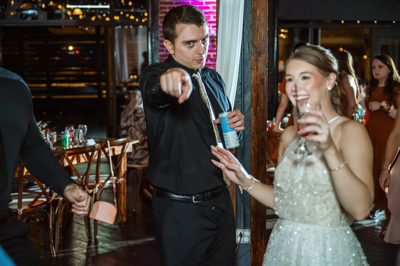 Man in black shirt points at the camera at a lively wedding reception while a bride smiles nearby.