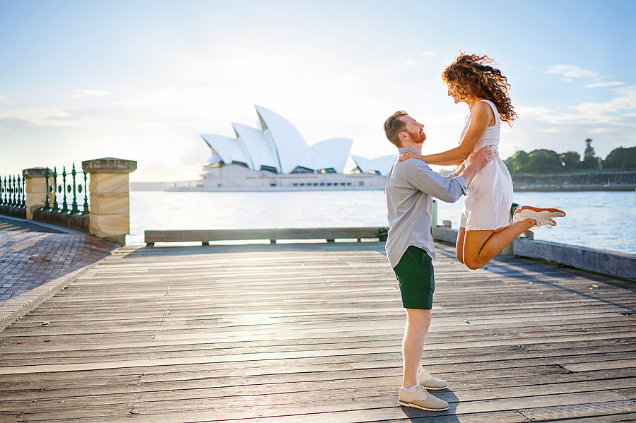 A photo of a man lifting a woman in a white dress into the air with the Sydney opera House in the background.