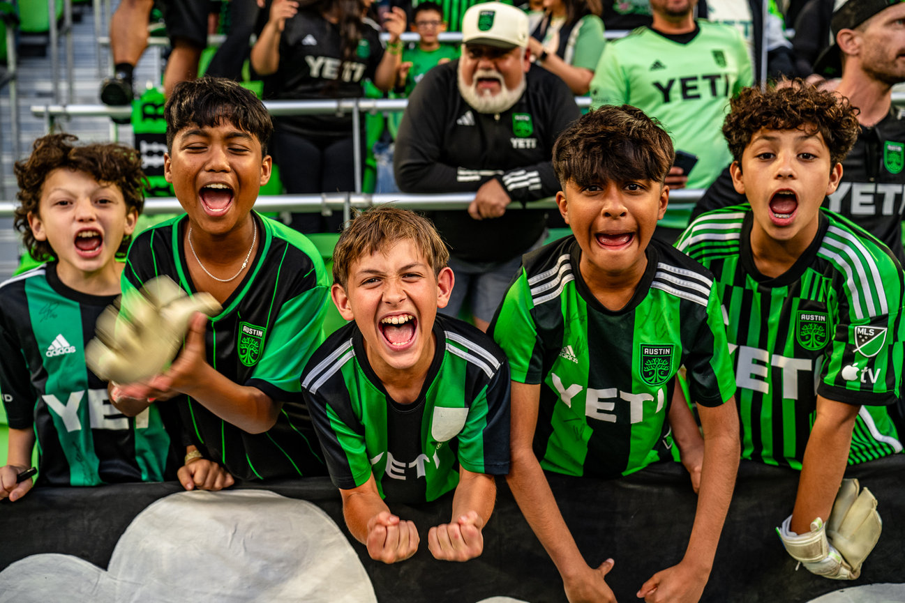 Five enthusiastic kids wearing Austin FC jerseys, cheering at an Austin FC soccer game, displaying excitement and team spirit.