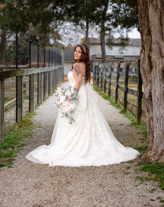 Bride smiling over her shoulder while holding a pastel bouquet along a rustic farm fence at her outdoor wedding captured by Gulf Coast Wedding photographer Dixon Creative Images