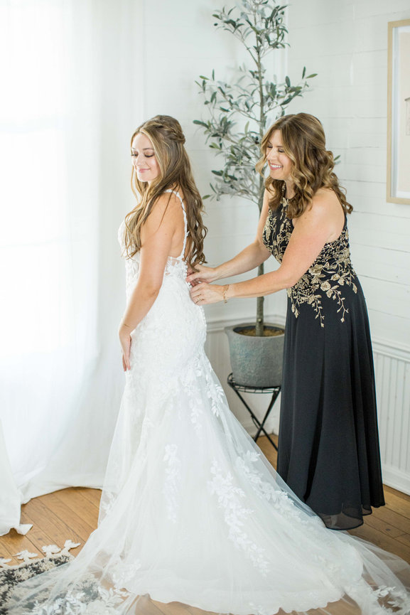 Mother of bride zipping brides dress while bride looks over her shoulder and smiles