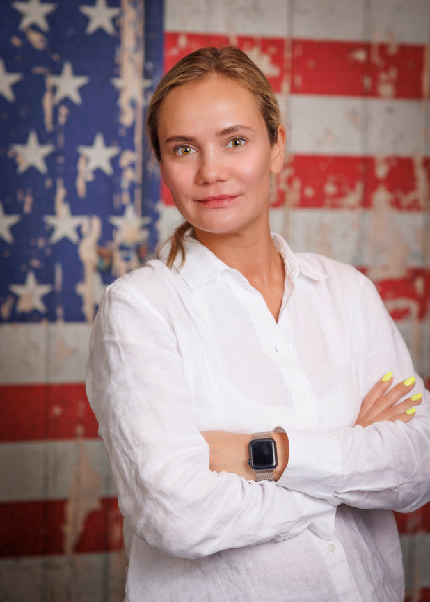 Woman in a white shirt with arms crossed, standing in front of a weathered American flag backdrop.