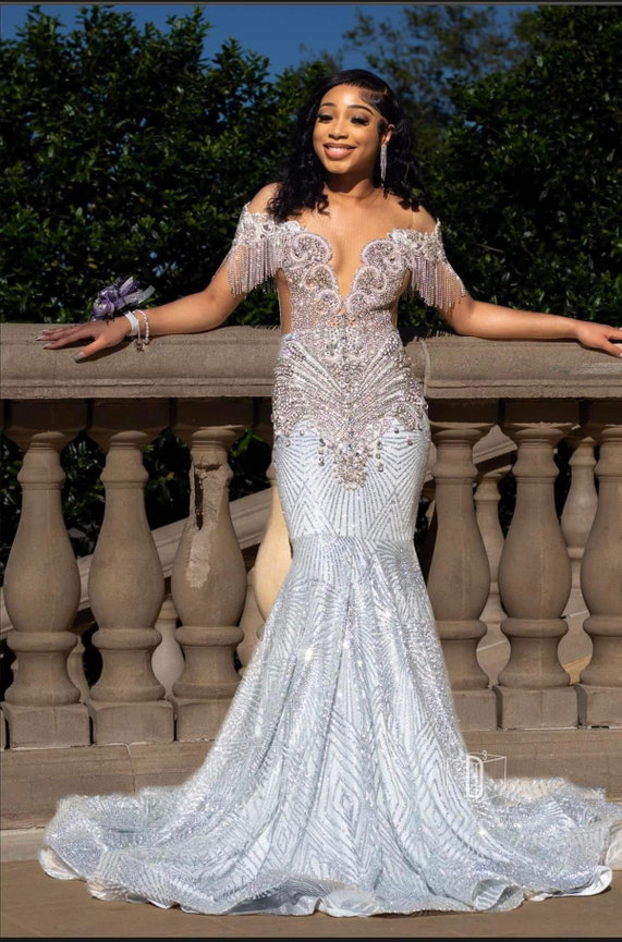 Woman in an elegant, silver off-shoulder gown with intricate beadwork, standing by a stone balustrade with greenery behind.