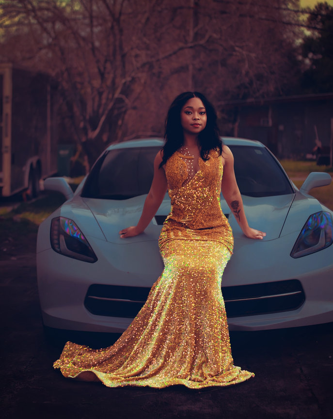 Woman in a shimmering gold dress sitting on a light blue sports car, with a wooded background.