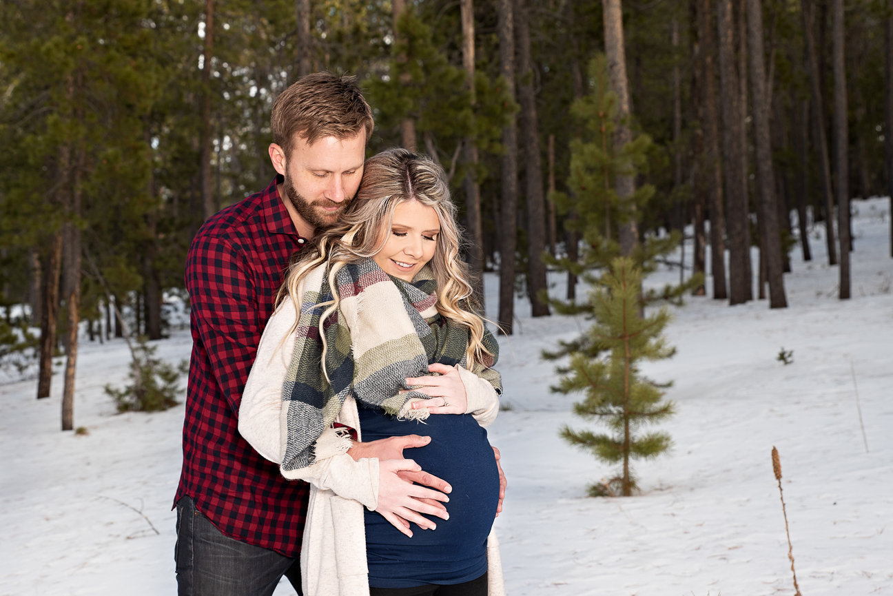 Couple embracing in snowy forest, with woman holding a pregnant belly, in cozy clothing.