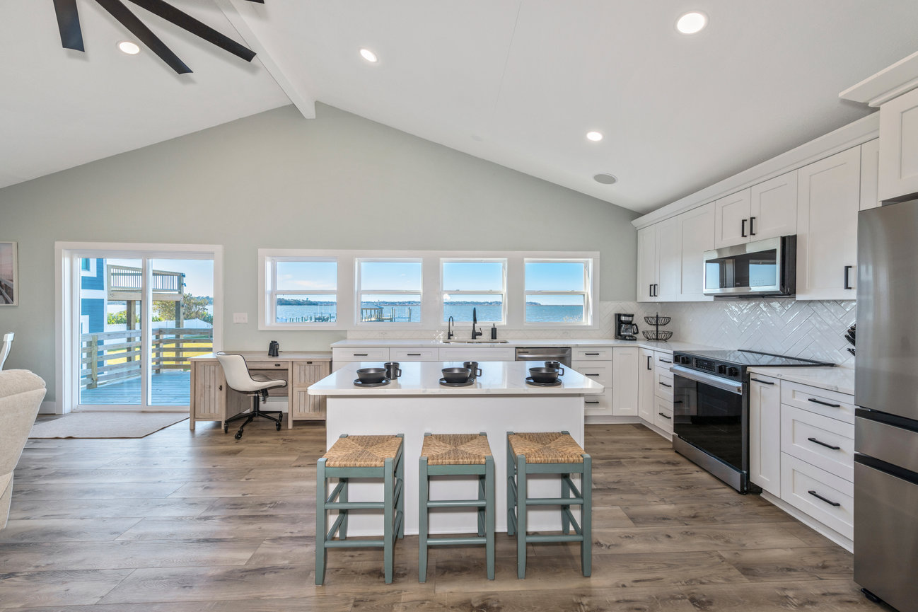 Modern kitchen with white cabinets, center island with stools, stainless steel appliances, and large windows with ocean view.