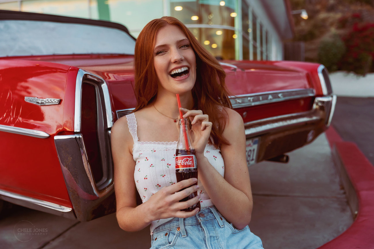 A REDHEAD WEARING A SUMMER TANK TOP AND SHORTS AND SKATES IS SITTING AGAINST A VINTAGE CAR DRINKING A COCA COLA AND LAUGHING.
