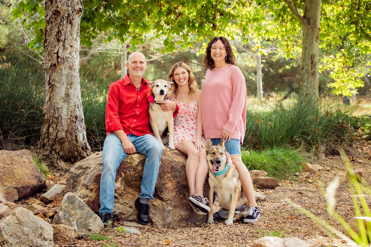 A HAPPY FAMILY WITH THEIR TWO DOGS IN A PARK HUGGING AND LAUGHING TOGETHER.