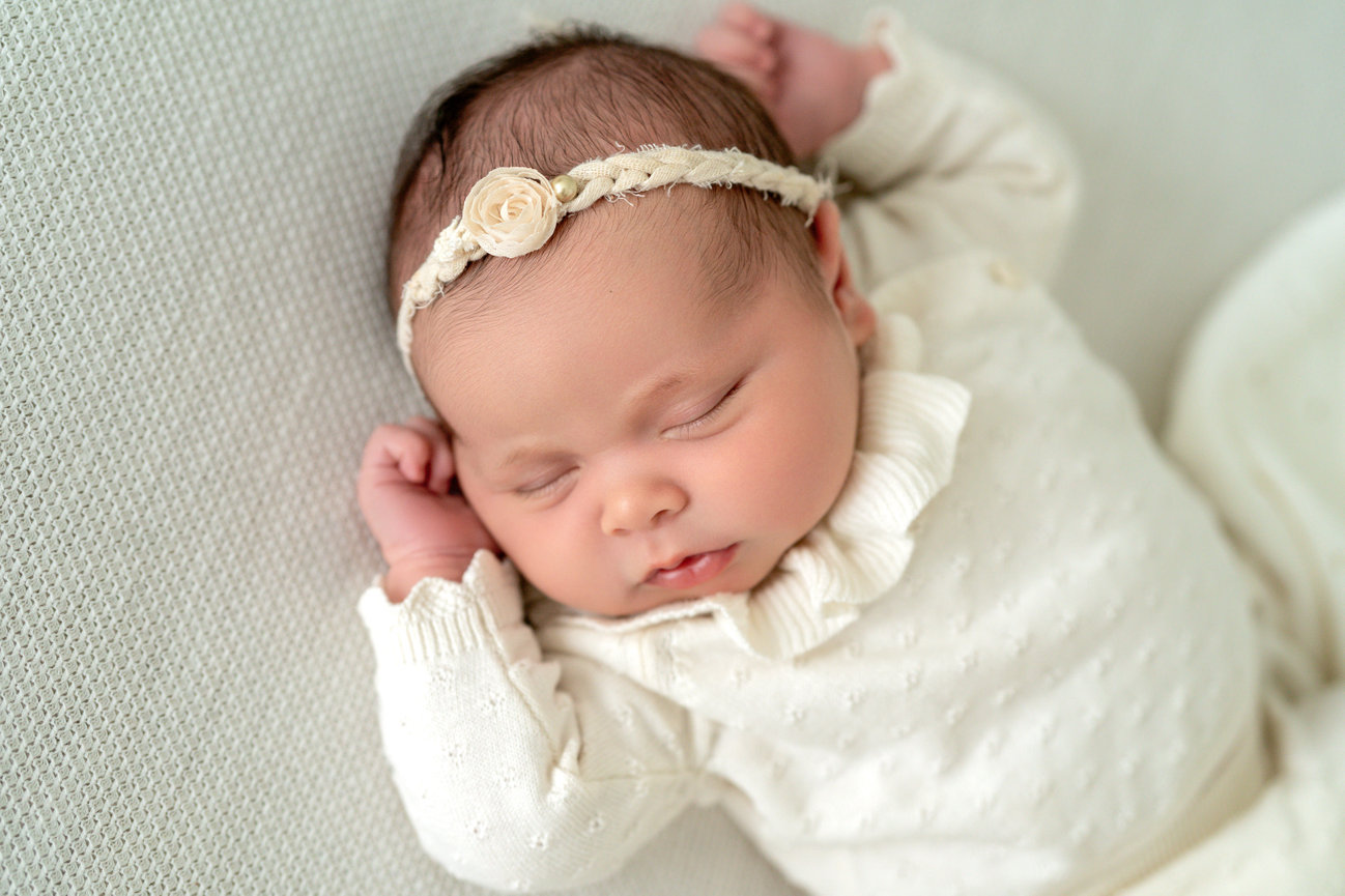 Sleeping newborn baby in a white outfit with floral headband, resting on a soft blanket
