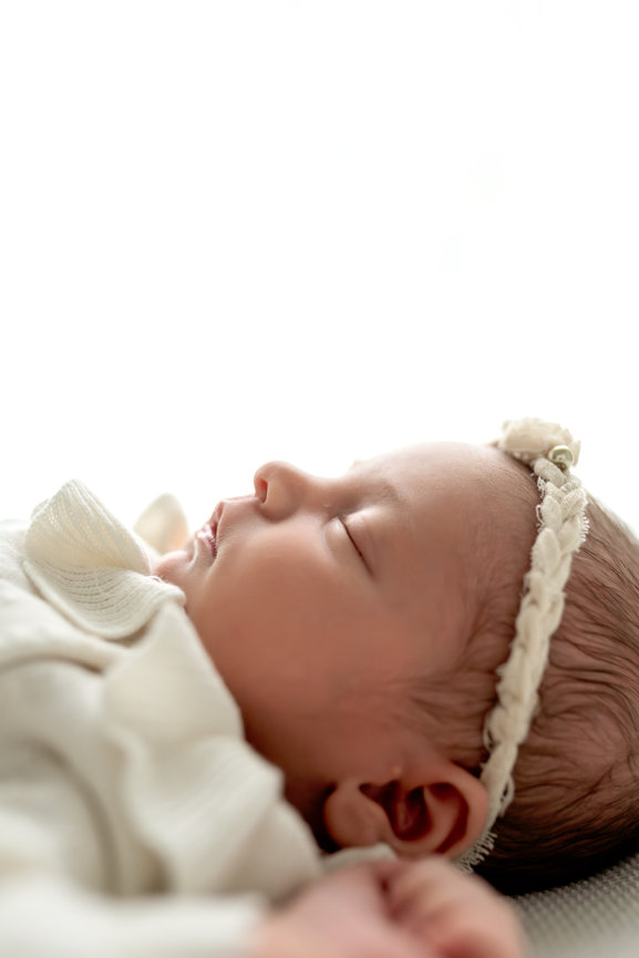 Sleeping newborn baby wearing a cream headband and soft white outfit on a bright background