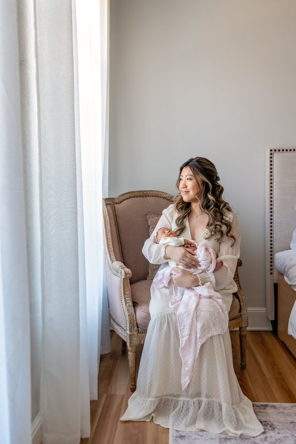 Mother holding a newborn baby in a softly lit nursery, sitting on a vintage chair near a window.
