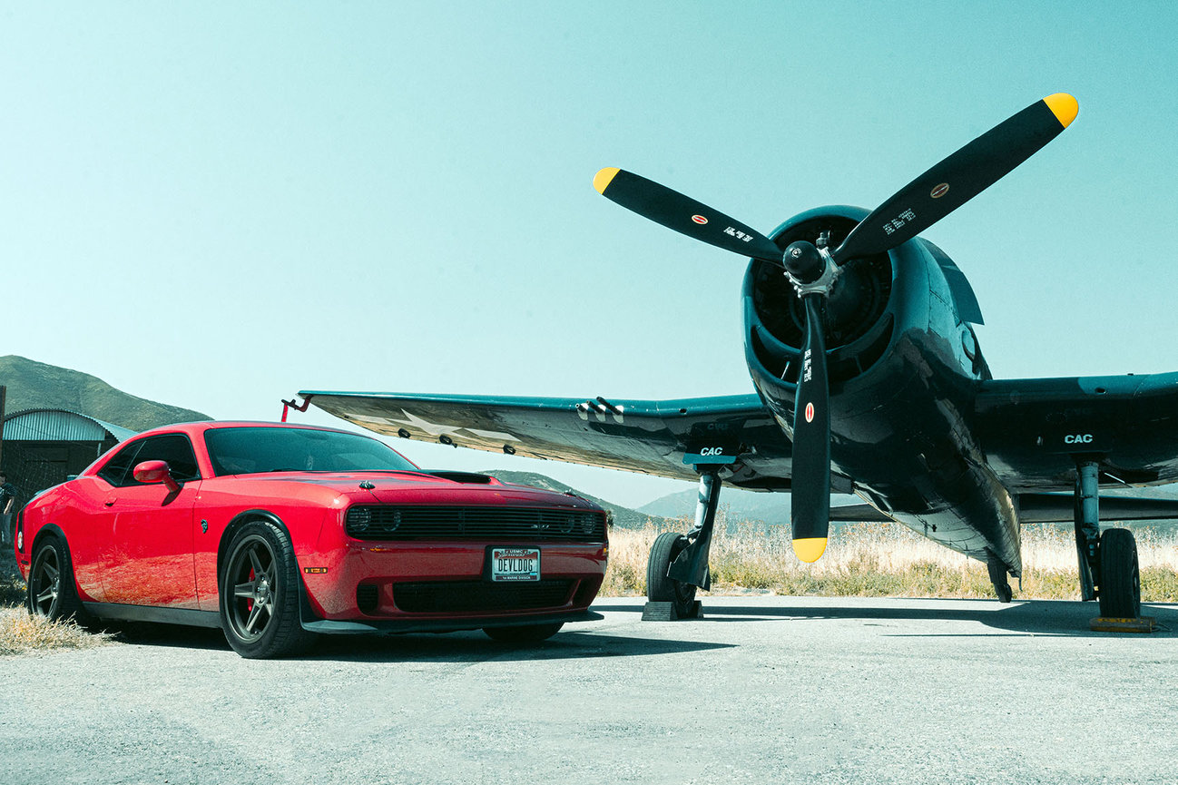 A red Dodge Challenger SRT Hellcat parked next to a vintage F6F Hellcat warplane on a sunny day in Utah. Photo shoot by Tre Nelson Photo.