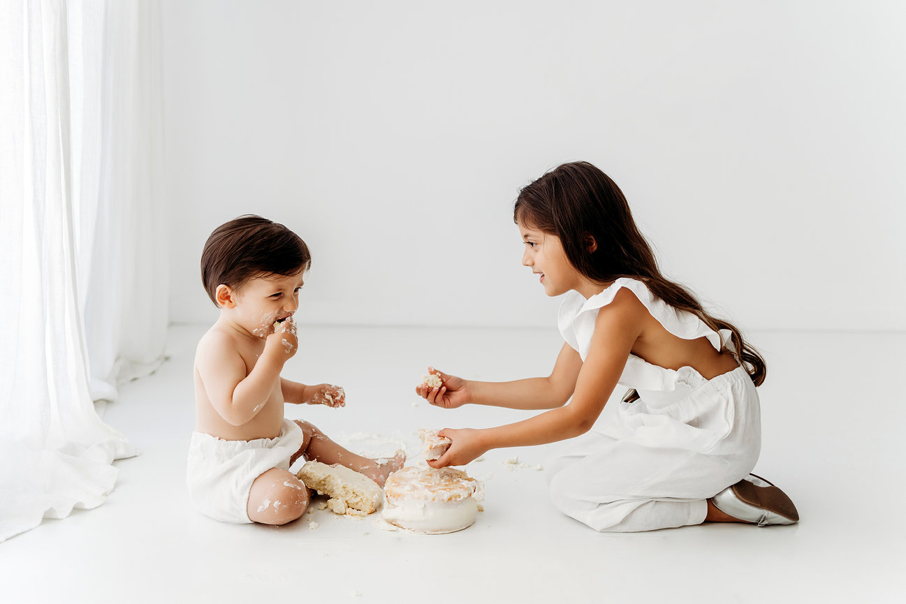 A child and a baby playing with cake on a white floor in a bright room.
