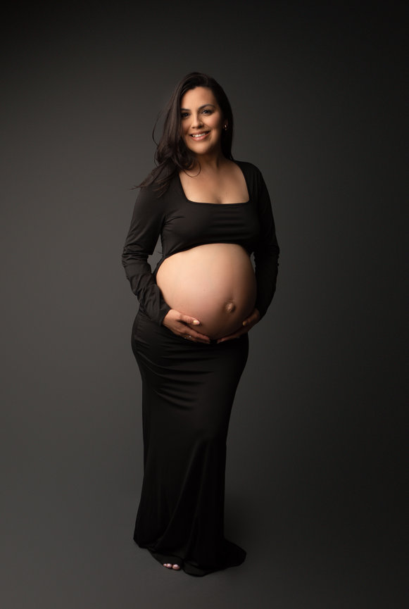 Pregnant woman in a long, black dress, smiling and holding her belly against a dark background.