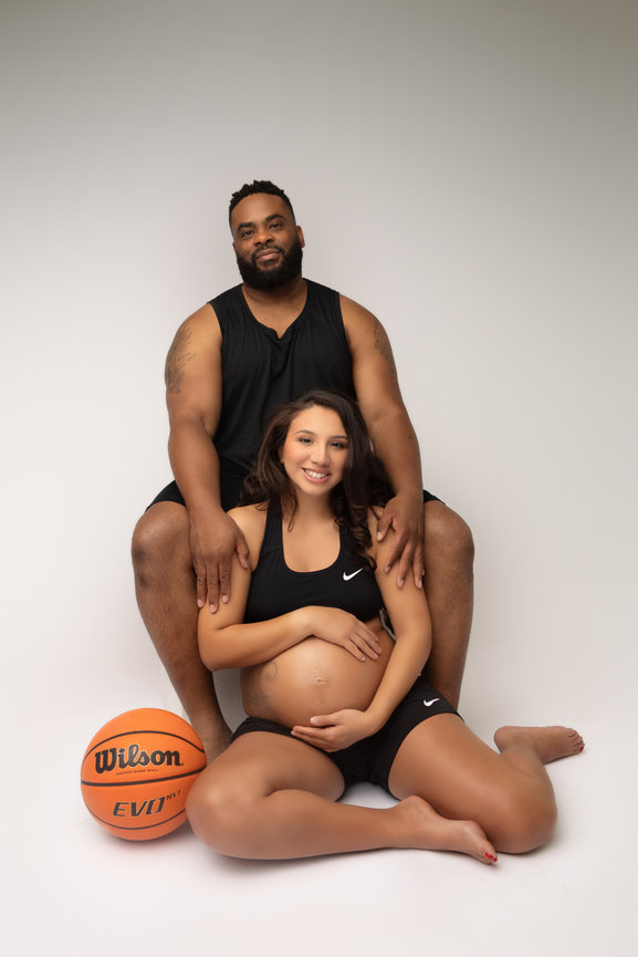 A couple in sportswear poses with a basketball. The pregnant woman sits in front, holding her belly, and the man sits behind her.