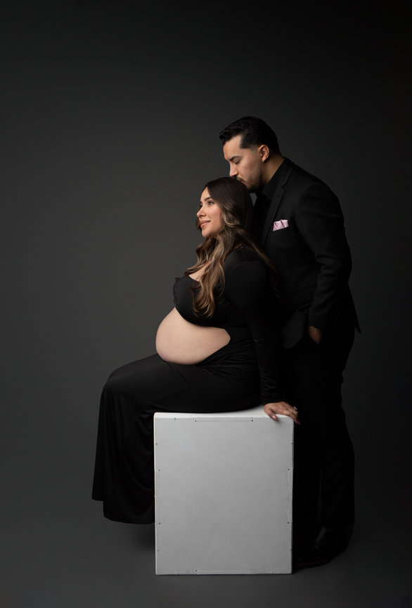 Pregnant woman seated on a white cube, elegantly dressed in black, with a man standing beside her in a black suit.