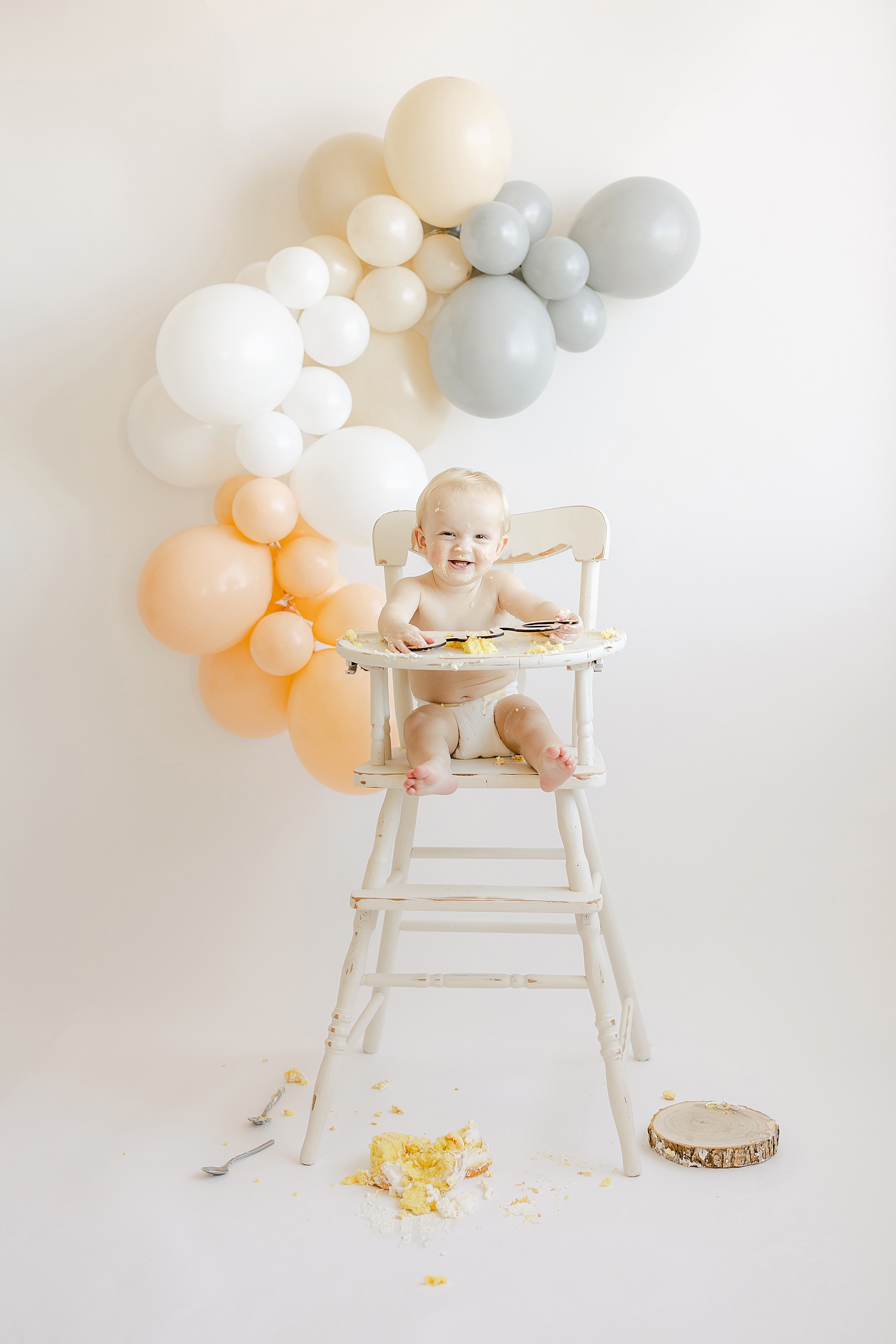 baby boy sitting in white high chair with white background and balloons eating yellow and white cake and smiling