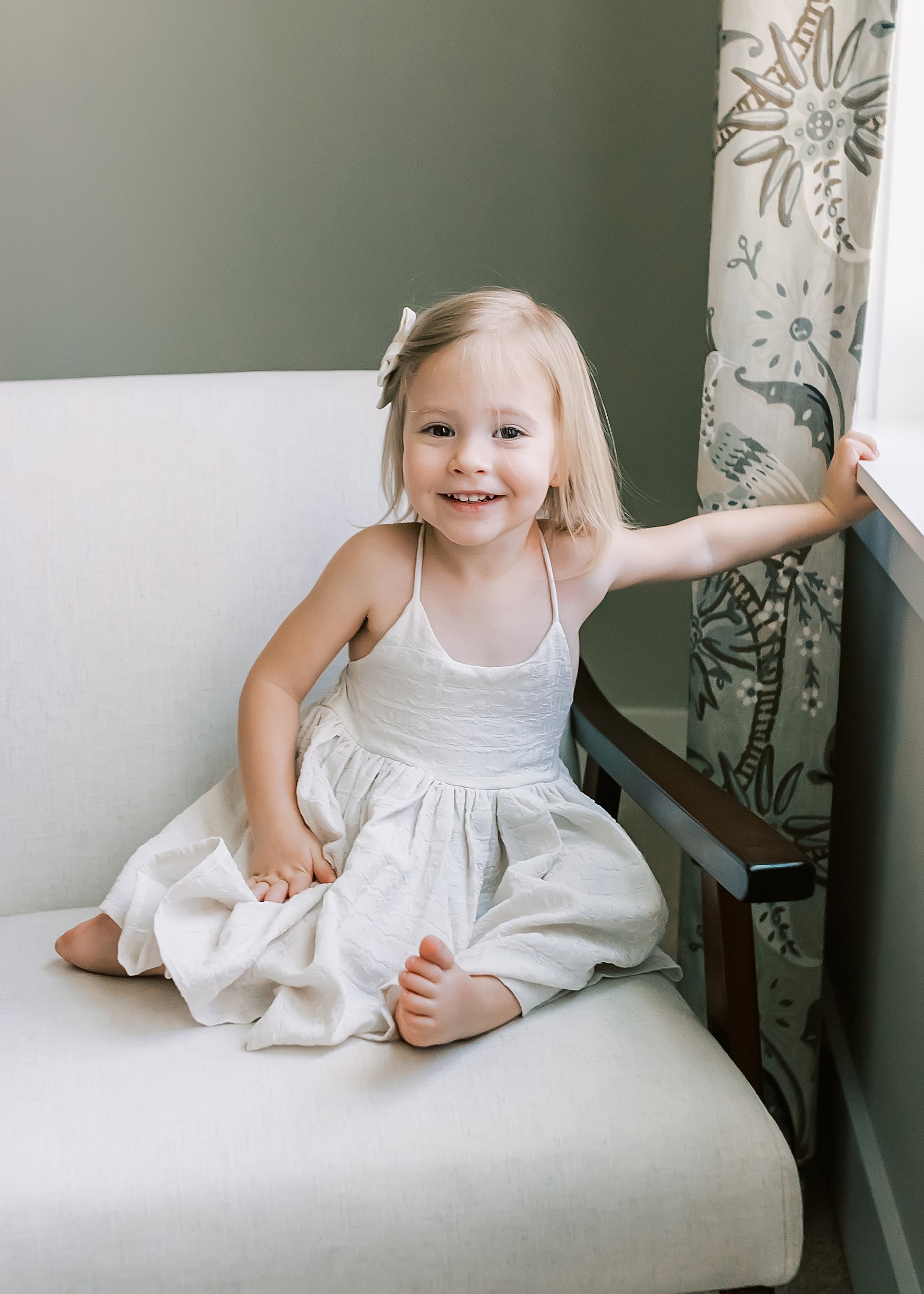 little girl dressed in neutral dress smiling at camera sitting on chair