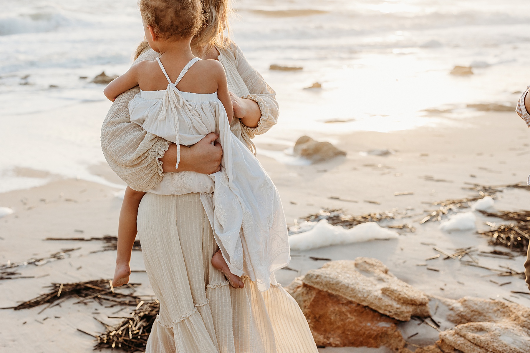 little girl wearing long white dress on the beach at sunrise being held by her mothers