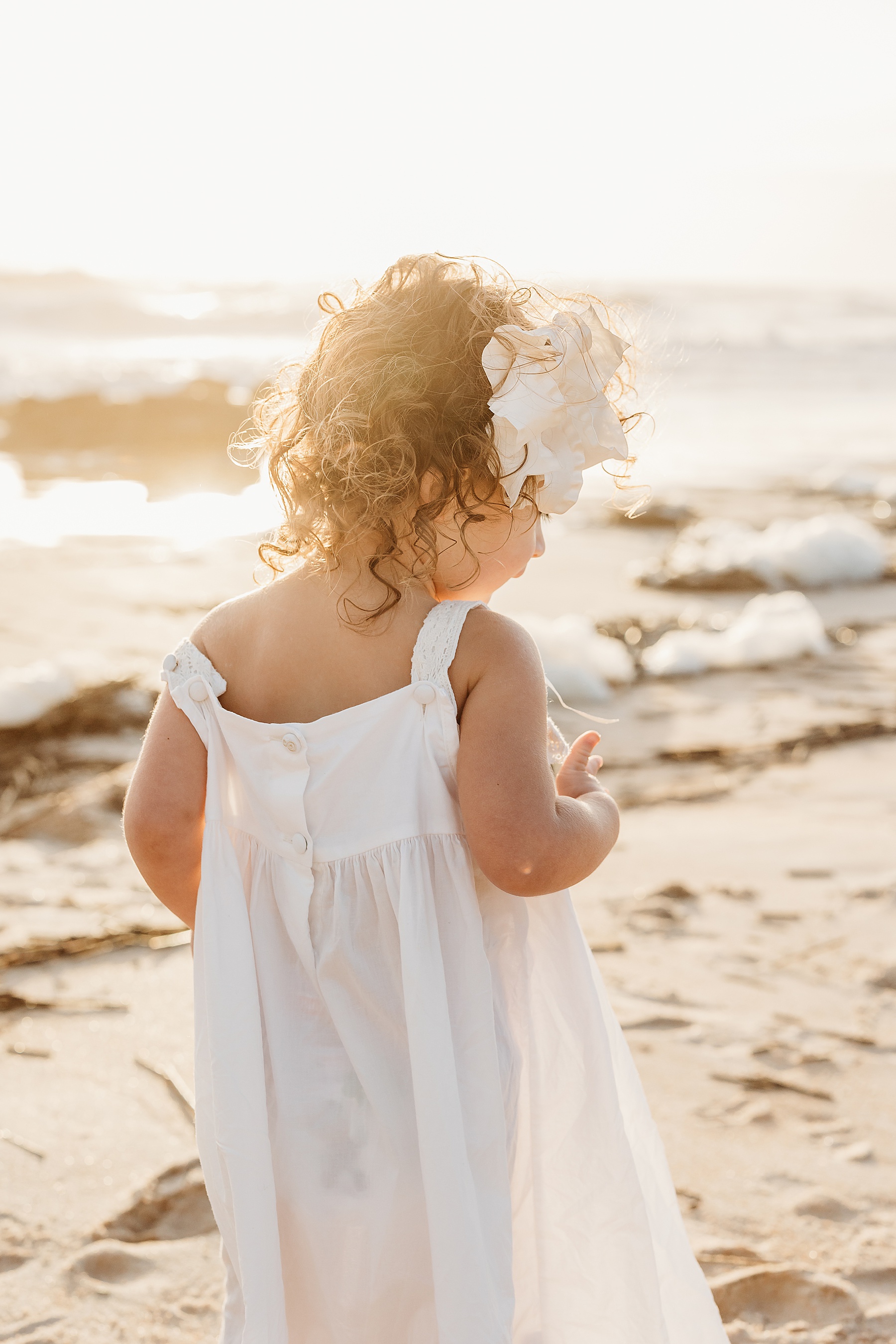 little girl in long white dress with big bow walking on the beach at sunrise