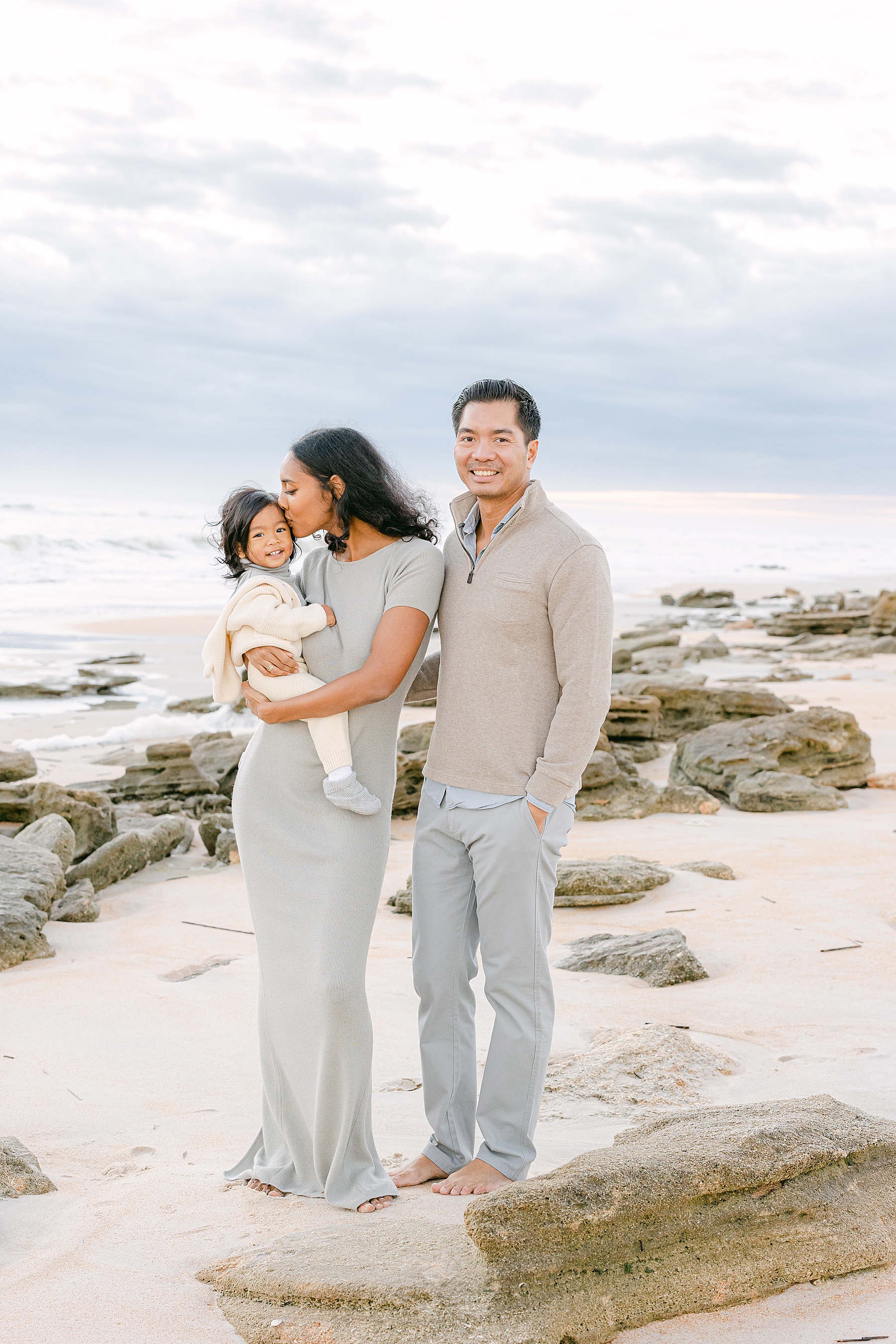 man and woman standing on the beach at sunrise wearing neutral color holding baby boy