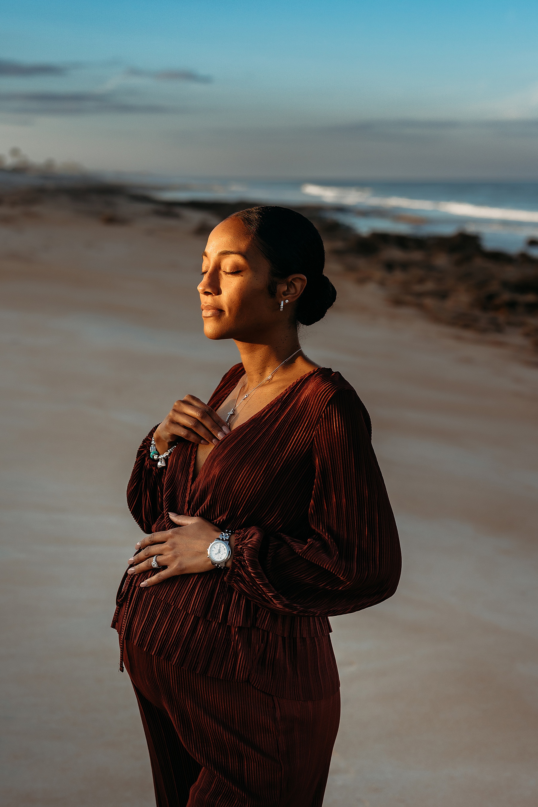 pregnant woman standing facing the golden sun on the beach with blue skies at sunset in saint augustine florida