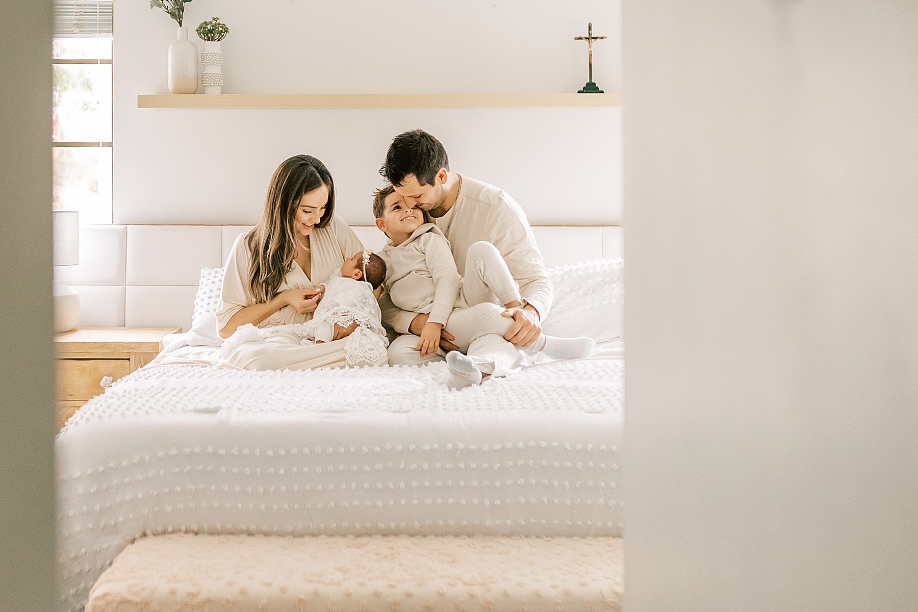 family dressed in tans and creams sitting on bed with white bedding holding baby girl