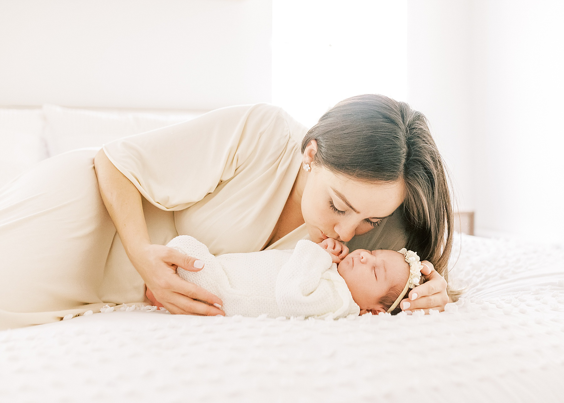 woman with long dark hair kissing newborn baby on white bed