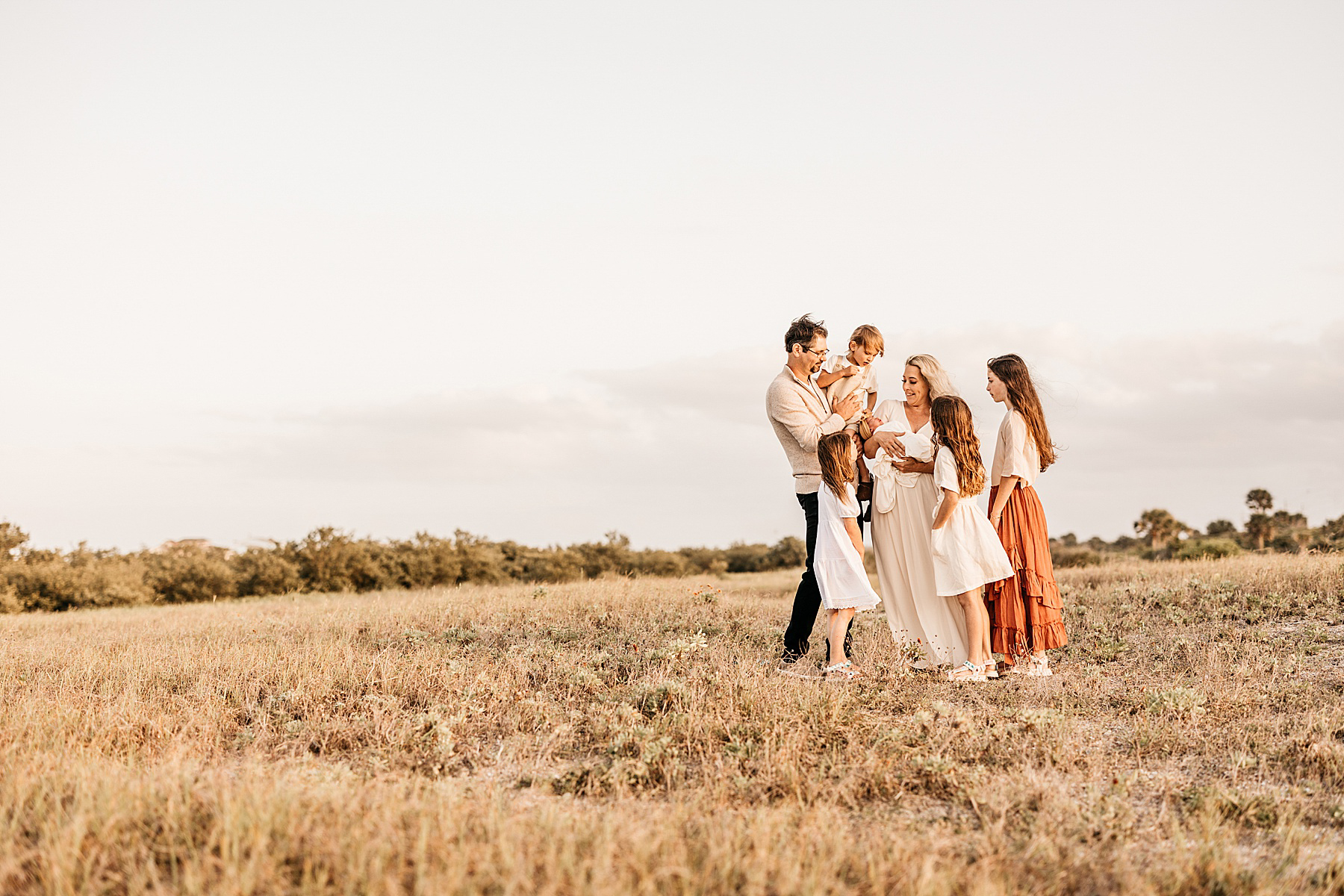 family wearing neutral colors standing in a grassy open field holding newborn baby boy