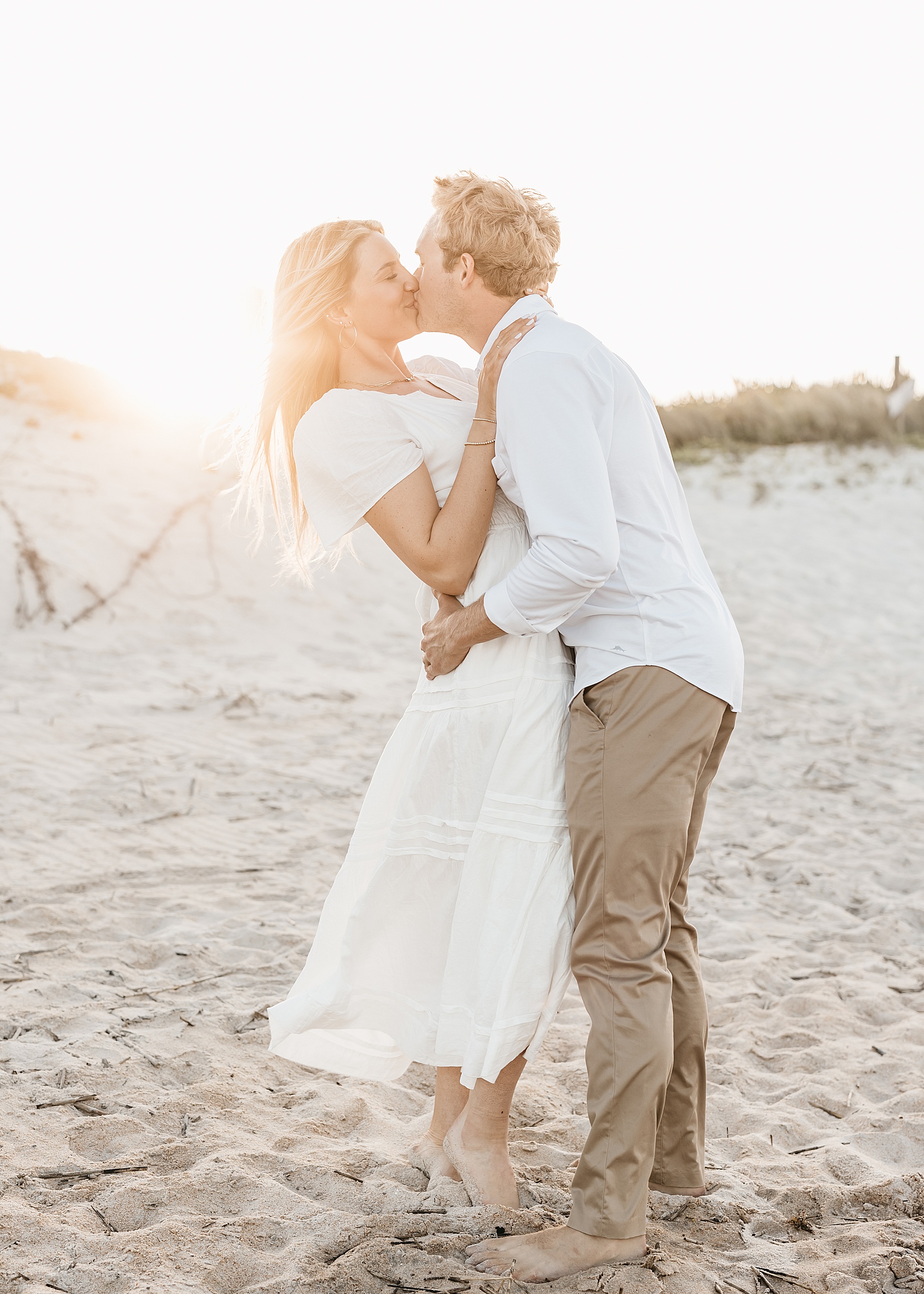 woman in white dress holding man in khaki pants with sun behind them on the sand